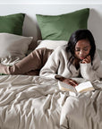 Woman reading a book on a bed with olive green and sand beige pillows.