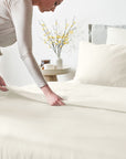 Person making a bed in a bright bedroom with white bedding and decorative elements.