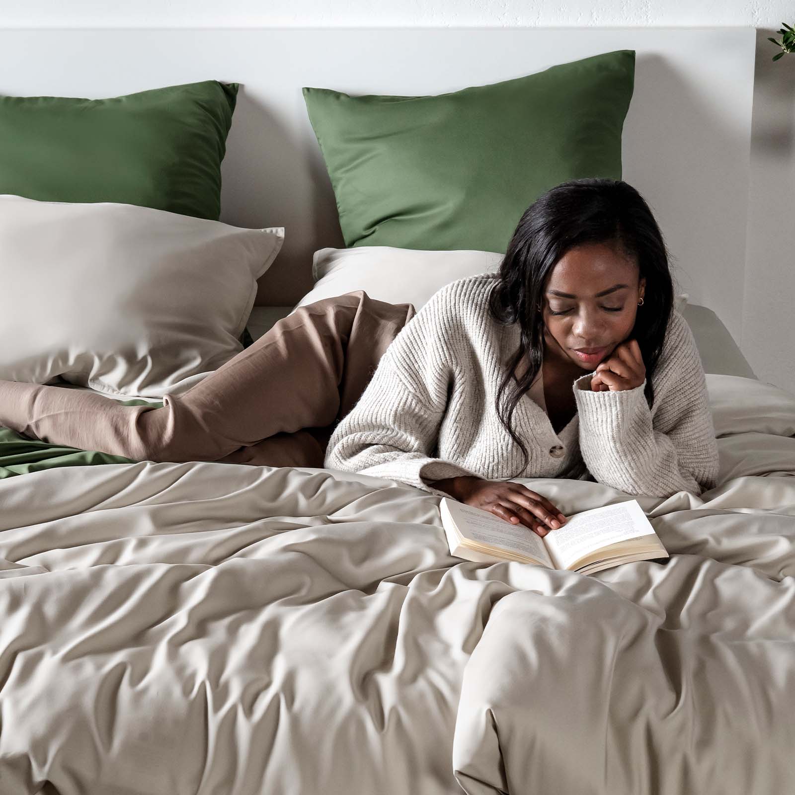 Woman reading a book on a bed with olive green and sand beige pillows.