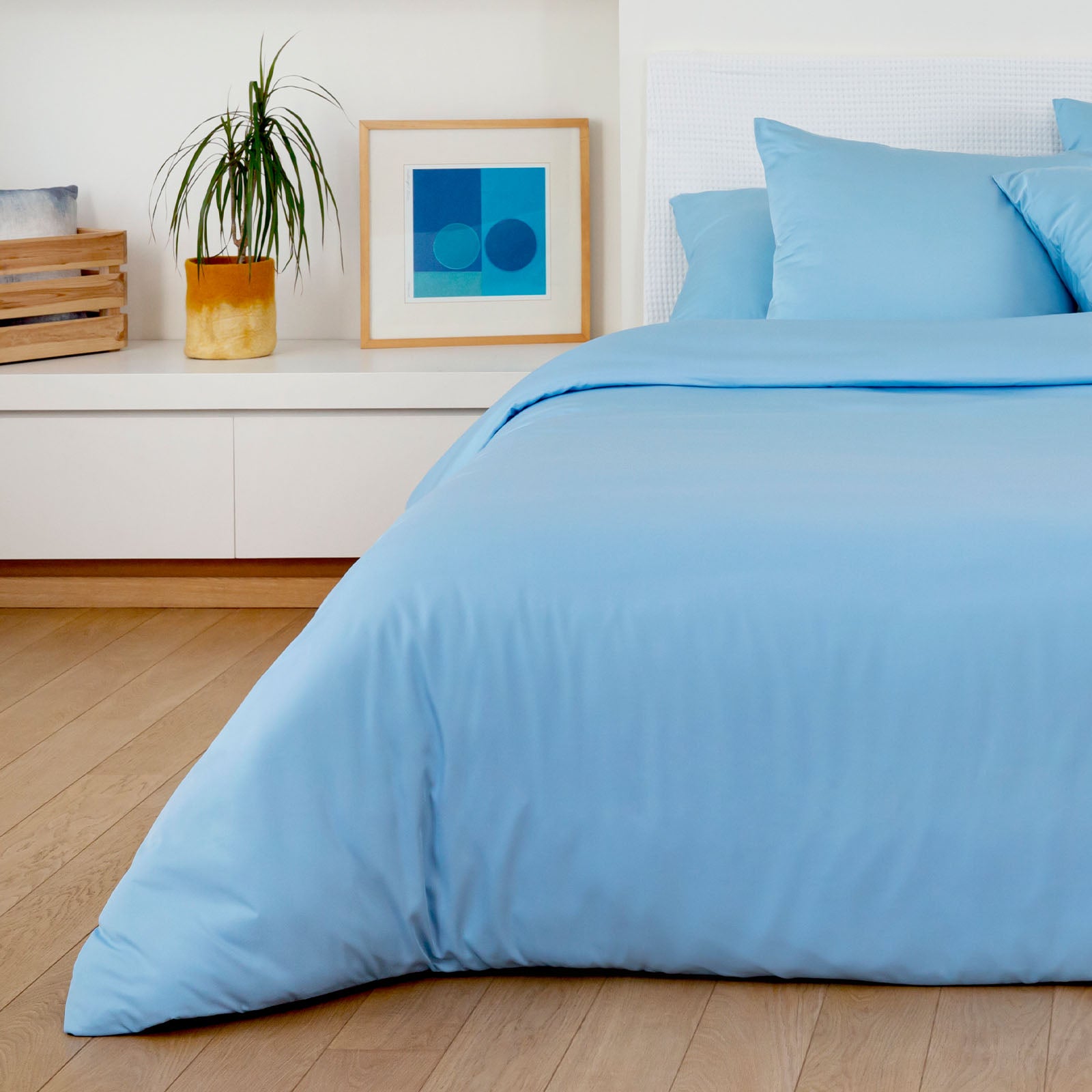 Blue bedding set on a bed in a room with a plant and artwork on a shelf.