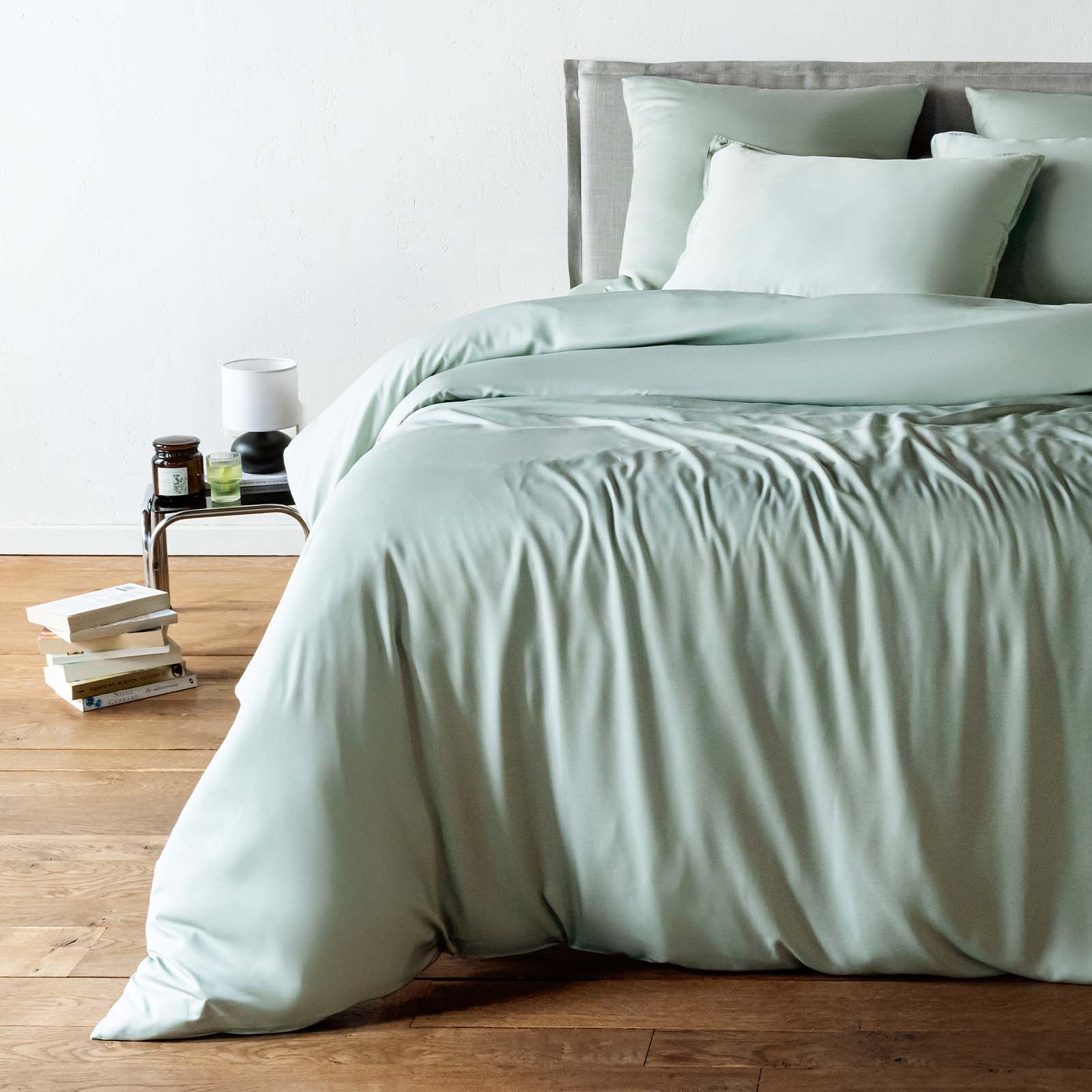 Bed with light green bedding in a room with a wooden floor and a nightstand.