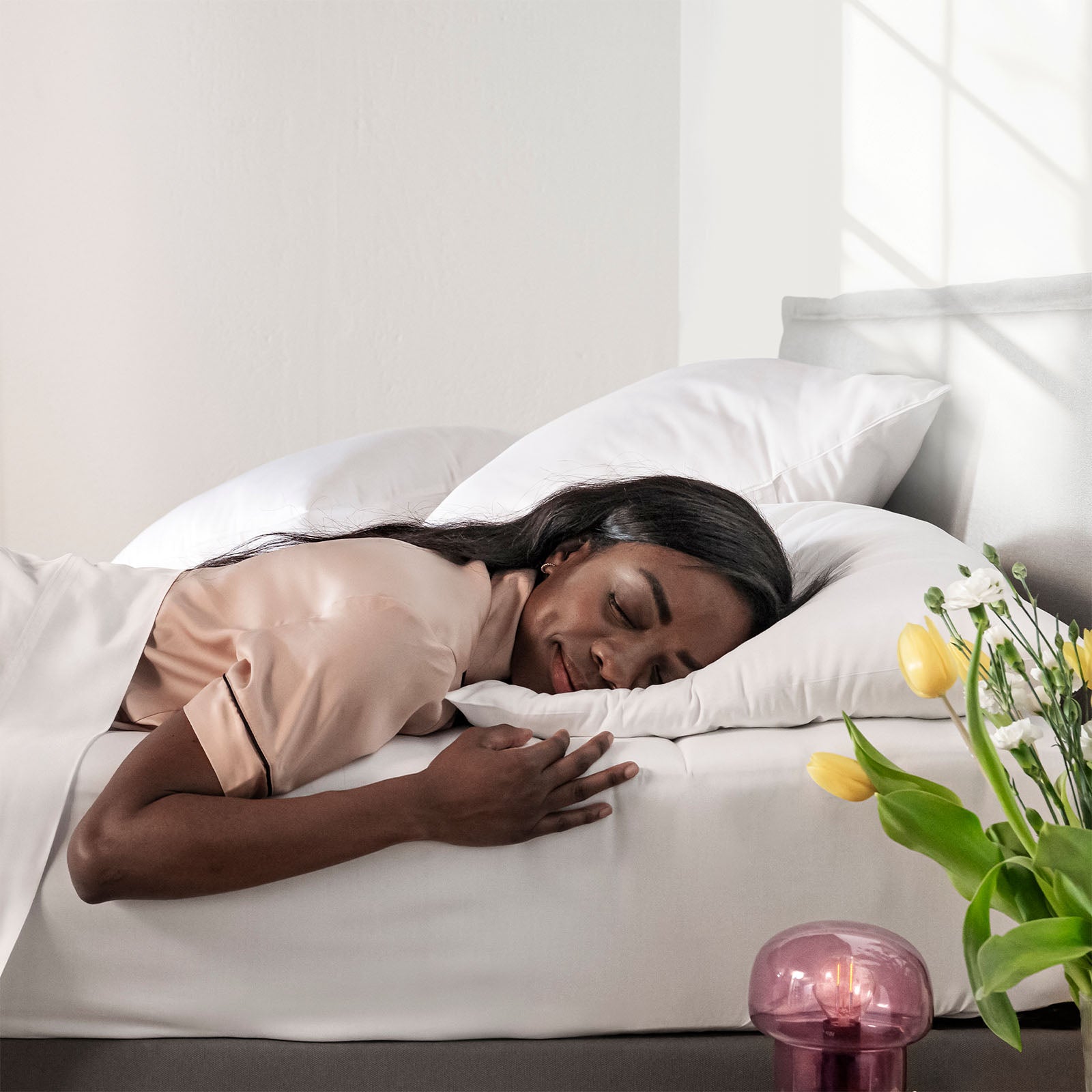 Woman sleeping peacefully in a bed with white bedding and a vase of flowers on the nightstand.