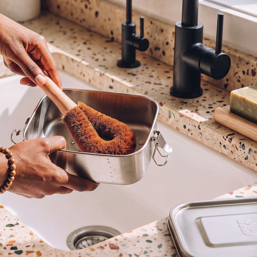 A plastic-free dish cleaning brush with a wooden handle is being held over a kitchen sink by two hands, while washing a stainless steel lunch box. The sink has a speckled terrazzo countertop, black faucet handles, and a bar of soap on a wooden block.